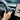 Hand gripping steering wheel while using turn signal lever stalk on Tesla Model 3 interior.